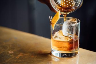 Pouring an Old Fashioned over ice in a clean rocks glass with orange garnish